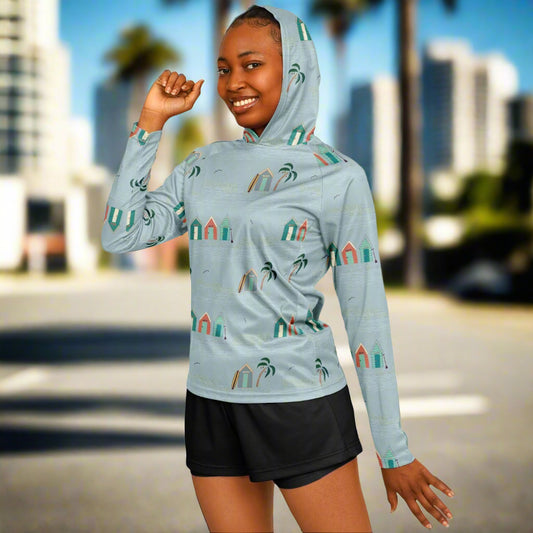 A woman wearing a long-sleeved sun hoodie with a pattern of beach huts, in a light blue colour.